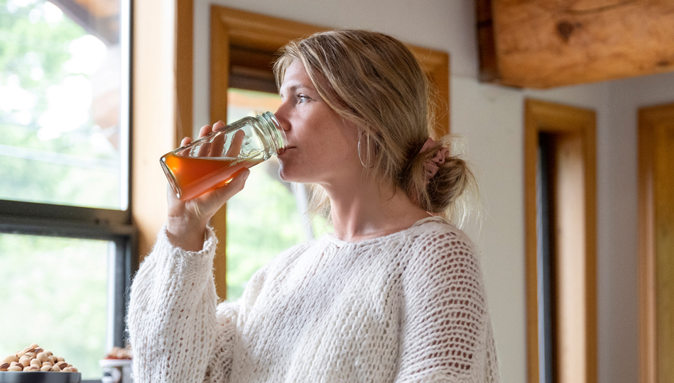 A woman gazes out the window while drinking a glass of kombucha.