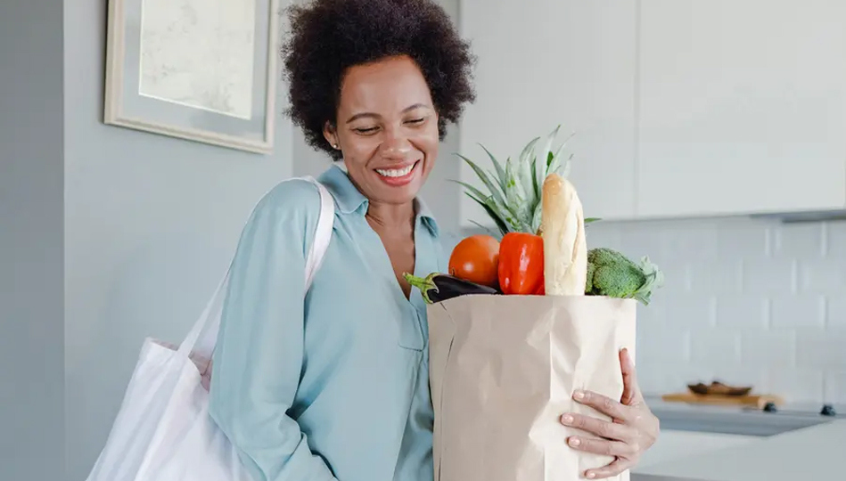 A woman carries a grocery bag full of fresh produce into her home.
